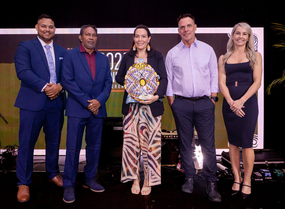 hree business professionals in matching formal attire standing on stage holding a hand-painted shield award during a national gala summit.