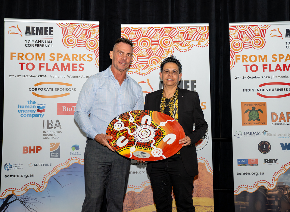 A smiling First Nations woman holding a brilliantly patterned hand-painted traditional shield award on stage at a formal evening gala.