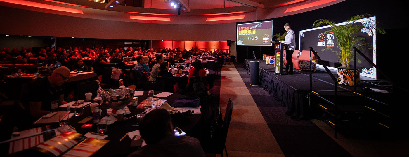 A wide shot of an active convention auditorium at an AEMEE conference, showing a CEO of AMEE, Jyi Lawton at the lectern addressing a seated audience during the Annual AEMEE Conference and Awards forum.