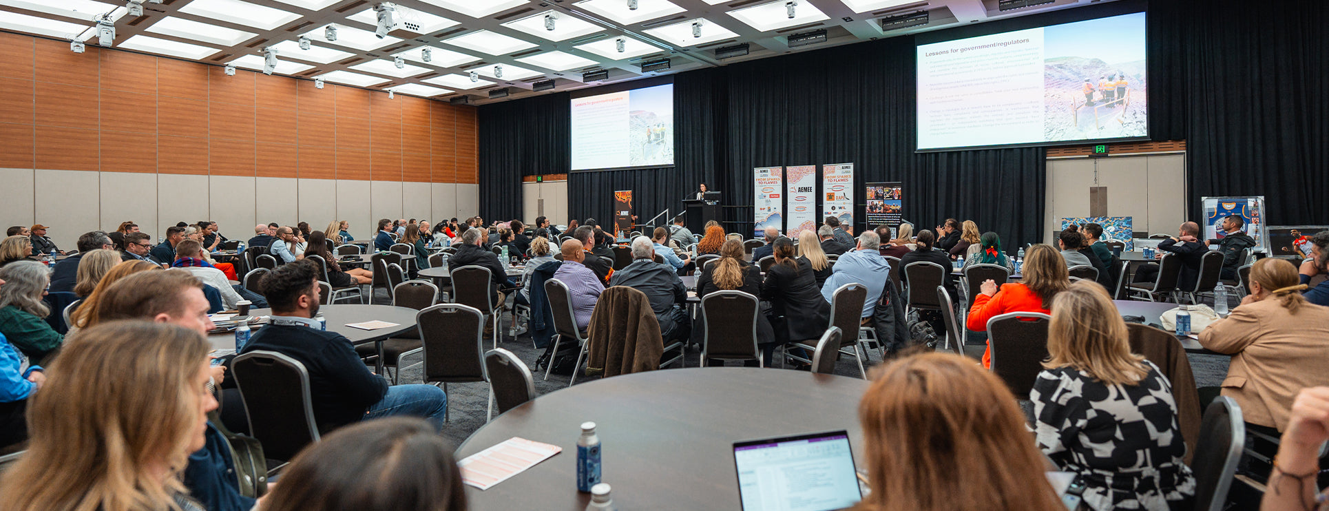 A wide shot of a crowded convention auditorium filled with delegates seated at round tables during an official AEMEE conference presentation, featuring a main speaker at the podium and large projector screens displaying data insights.