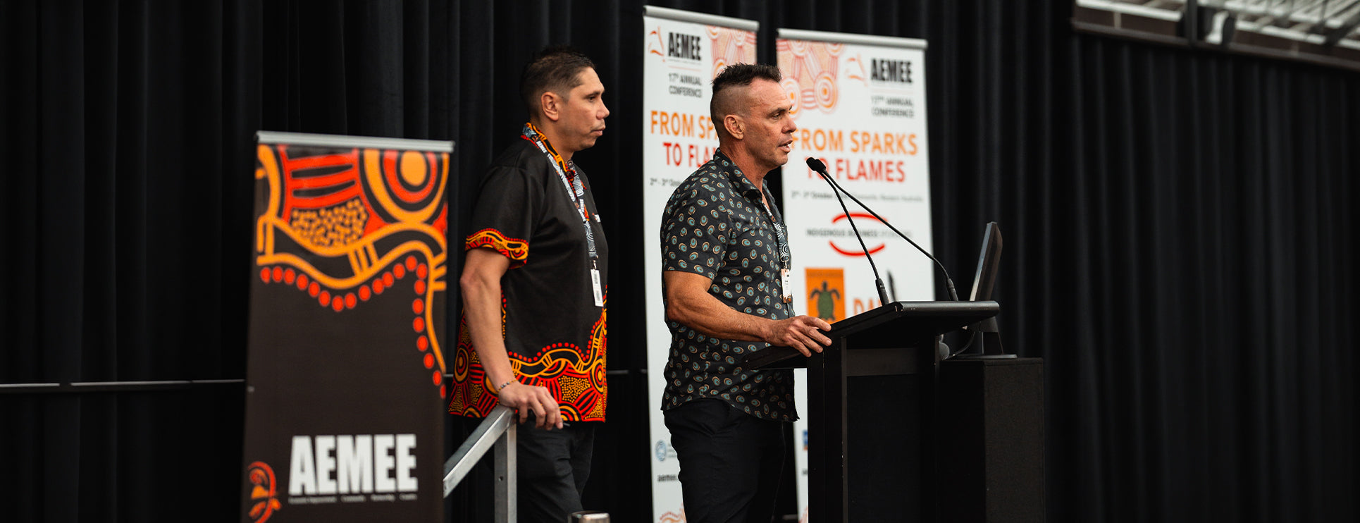 AEMEE Chief Executive Jyi Lawton stands alongside a presenter speaking at a corporate podium in front of 'From Sparks to Flames' conference banners during a national resources event.