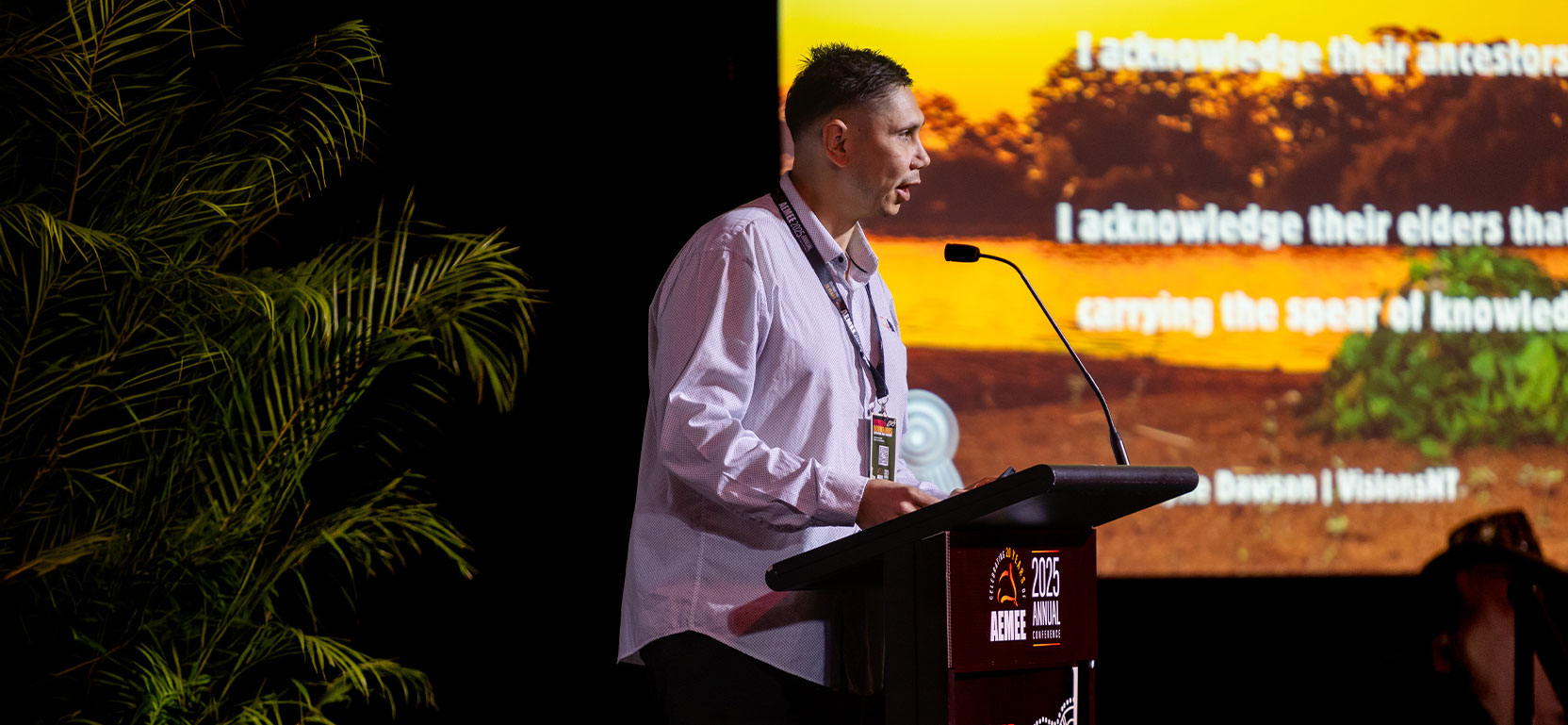 An AEMEE executive speaker delivering an industry report presentation at a podium in front of a large digital screen during a national resources sector summit.