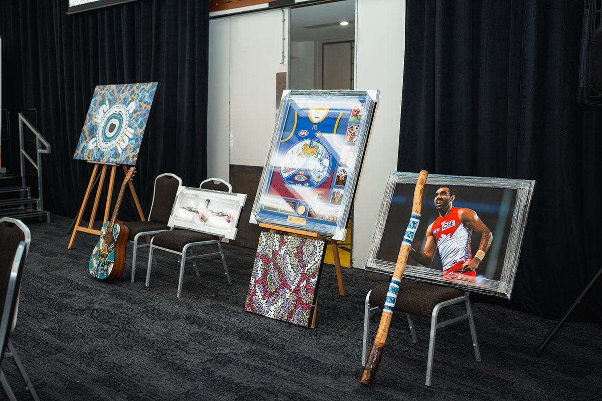 A collection of premium auction items on stage, including a hand-painted guitar, framed indigenous artwork prints, and a custom decorated wooden didgeridoo.