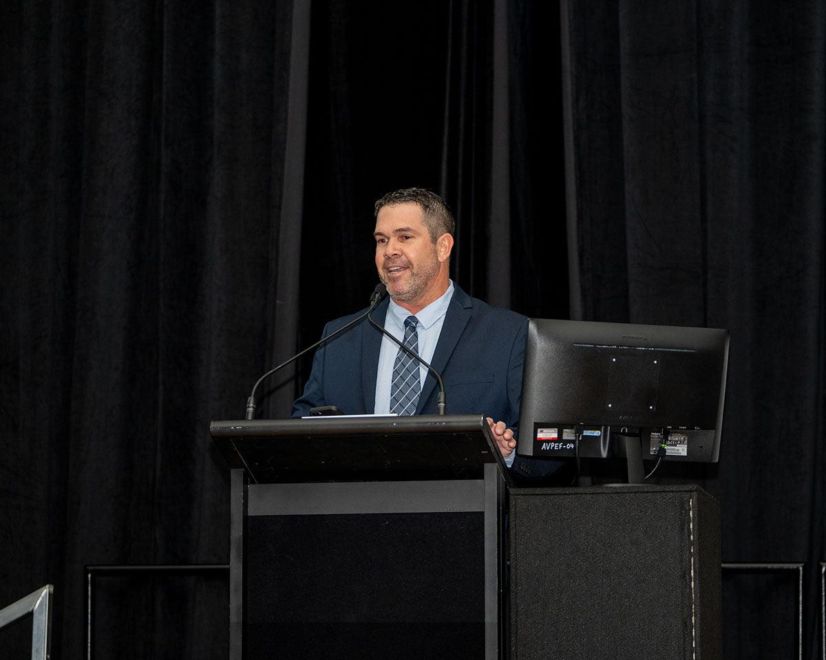 Brendan Bishop, Managing Director of First Enterprises, delivering an official address behind the podium on stage against a dark curtain backdrop.
