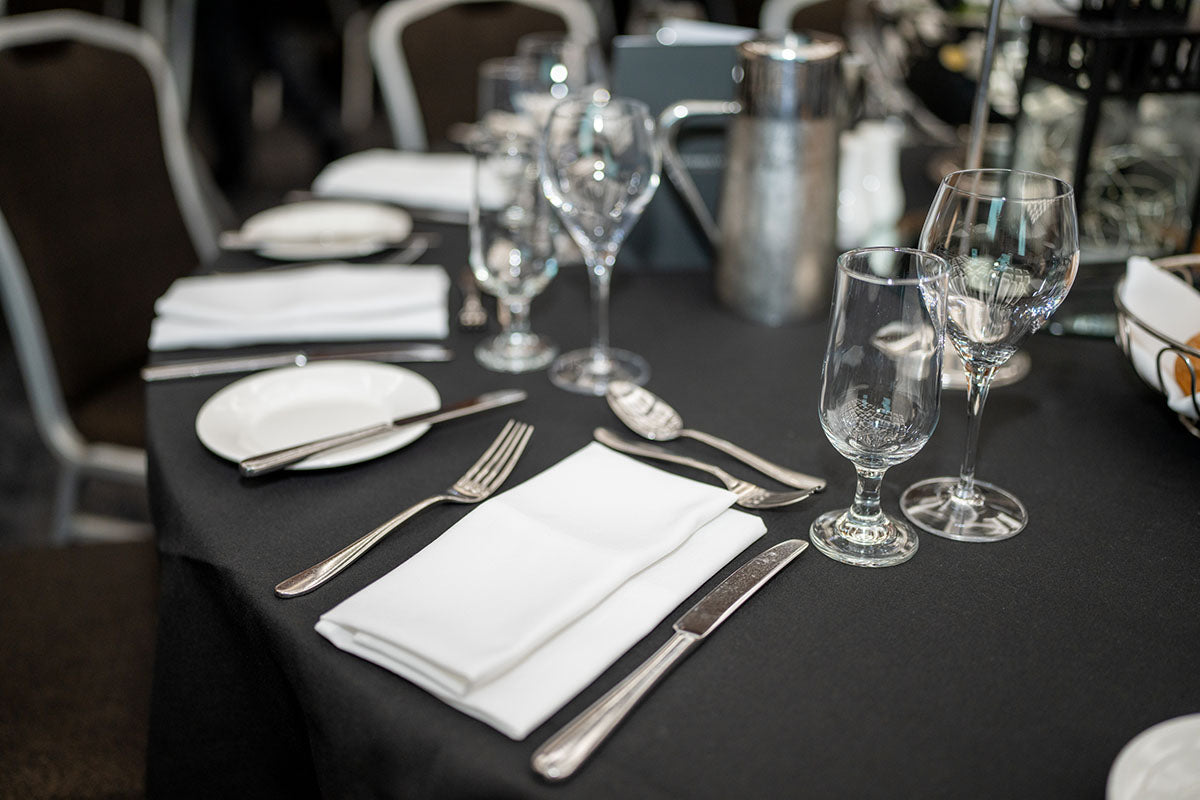 A formal banquet table setup for the awards dinner featuring clean black tablecloths, polished silverware, crisp white napkins, and elegant glassware.