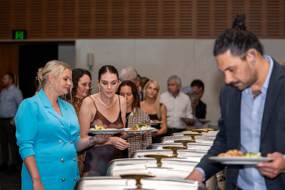 Conference attendees in formal dress wear holding plates and selecting dishes along the hot food buffet line.