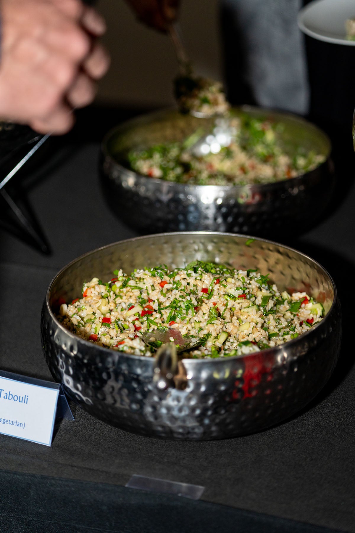 Close-up of a hand using tongs to serve fresh green grain salad from a large serving bowl at the banquet station.