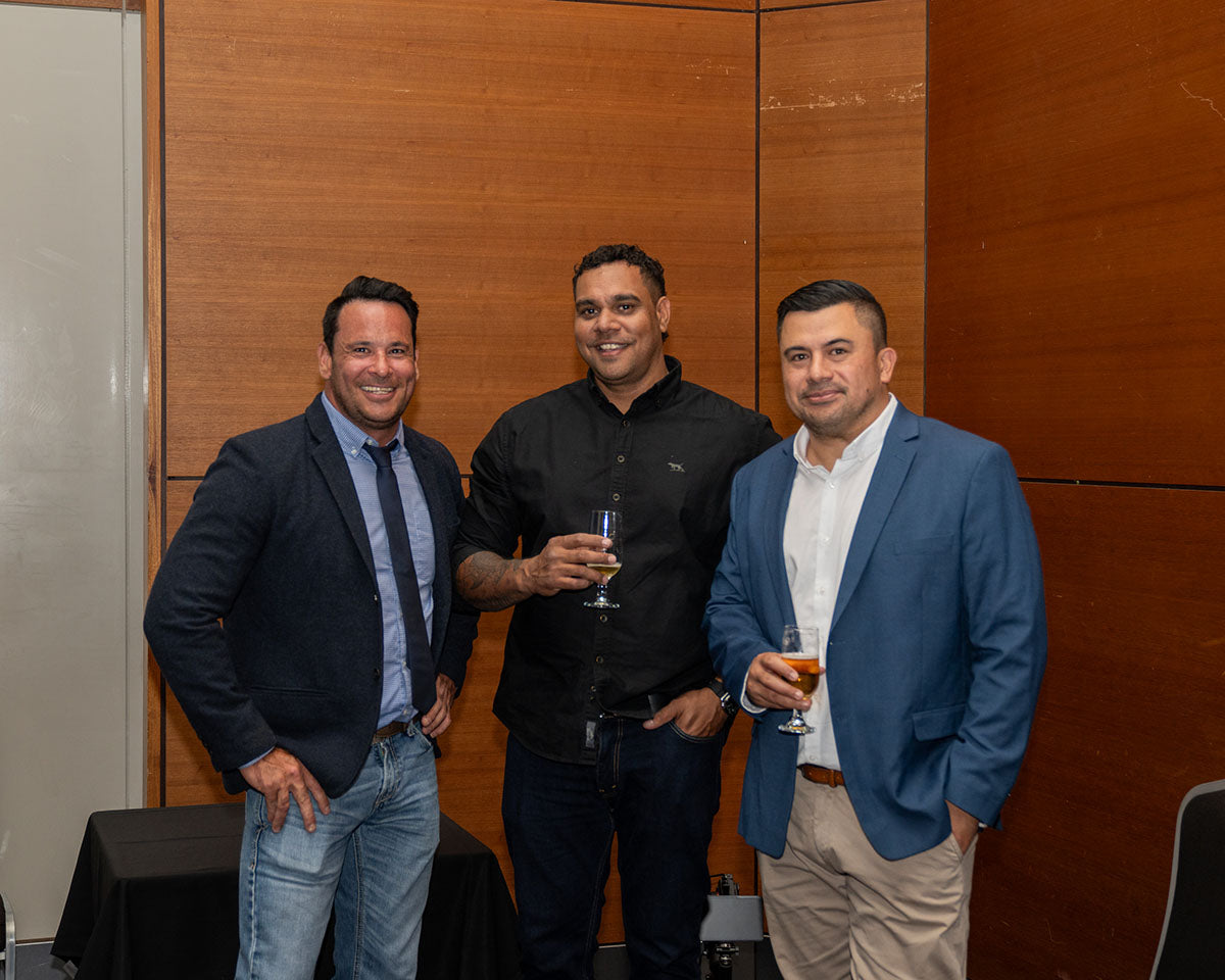 Three businessmen in smart casual jackets smiling together against a wood-paneled wall during the post-dinner networking block.
