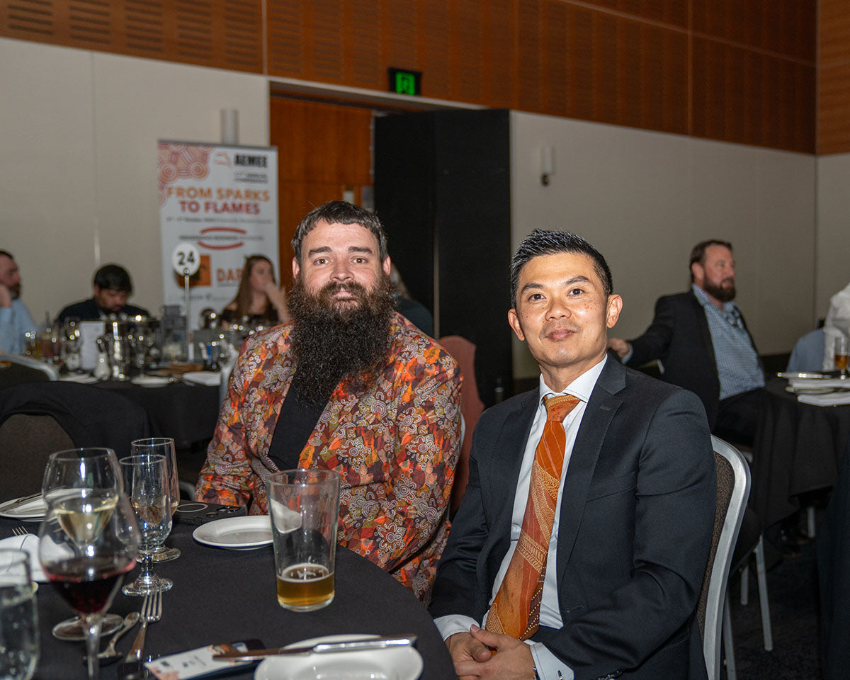 A male delegate with a long beard wearing a patterned orange blazer sitting with another businessman at a gala dinner table.