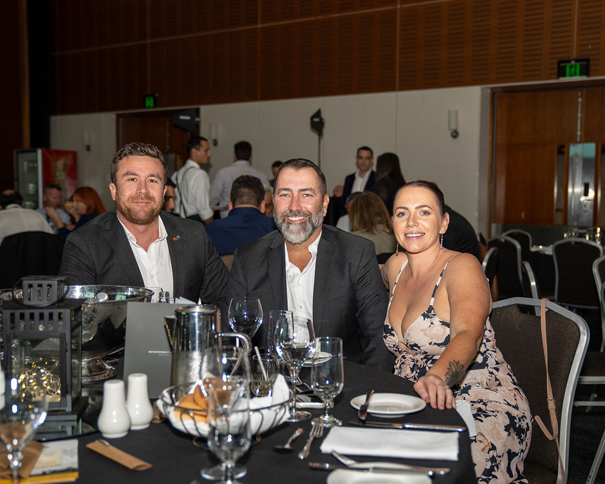 Three corporate representatives, two men in jackets and a woman in a floral dress, smiling from their banquet table during the awards evening.