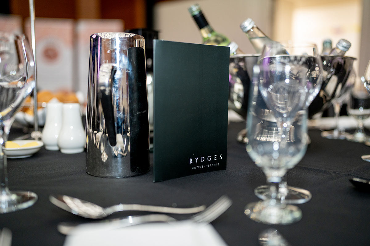 A close-up view of a dark green Rydges Hotels & Resorts table menu card standing next to a silver water pitcher on a set banquet table.