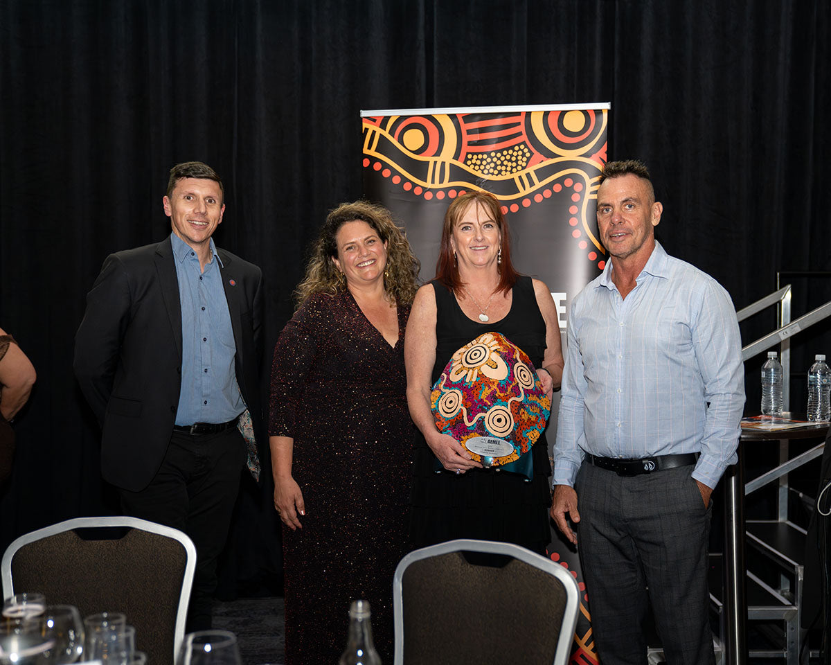 The winning team from Chevron posing proudly on stage with their large, custom hand-painted dot-art trophy shield after winning the 2024 Resource Sector Business of the Year award.
