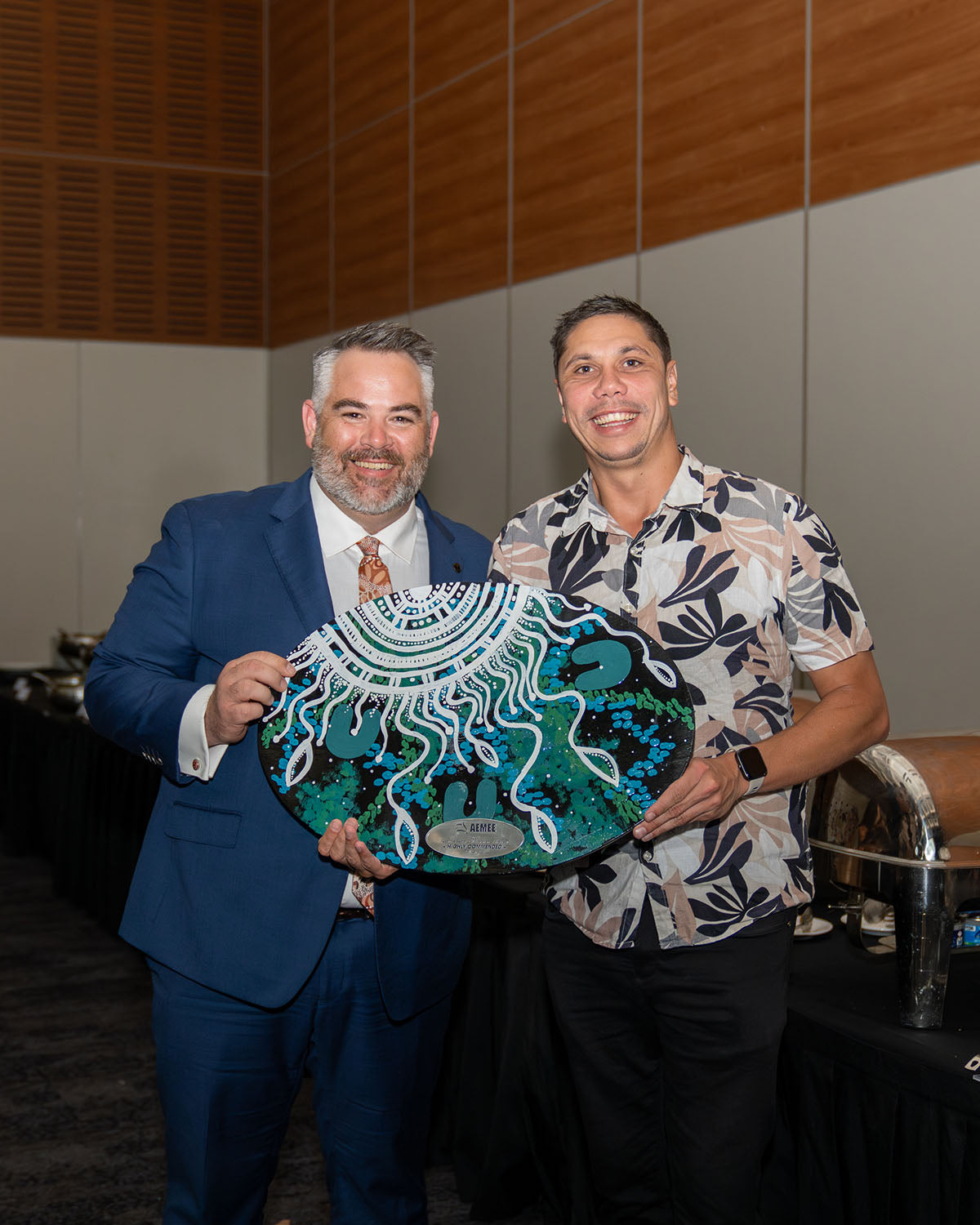 AEMEE CEO Jyi Lawton smiling broadly alongside an official corporate event sponsor representative who is holding up a custom green and blue hand-painted oval trophy plaque before presenting it to a category winner.
