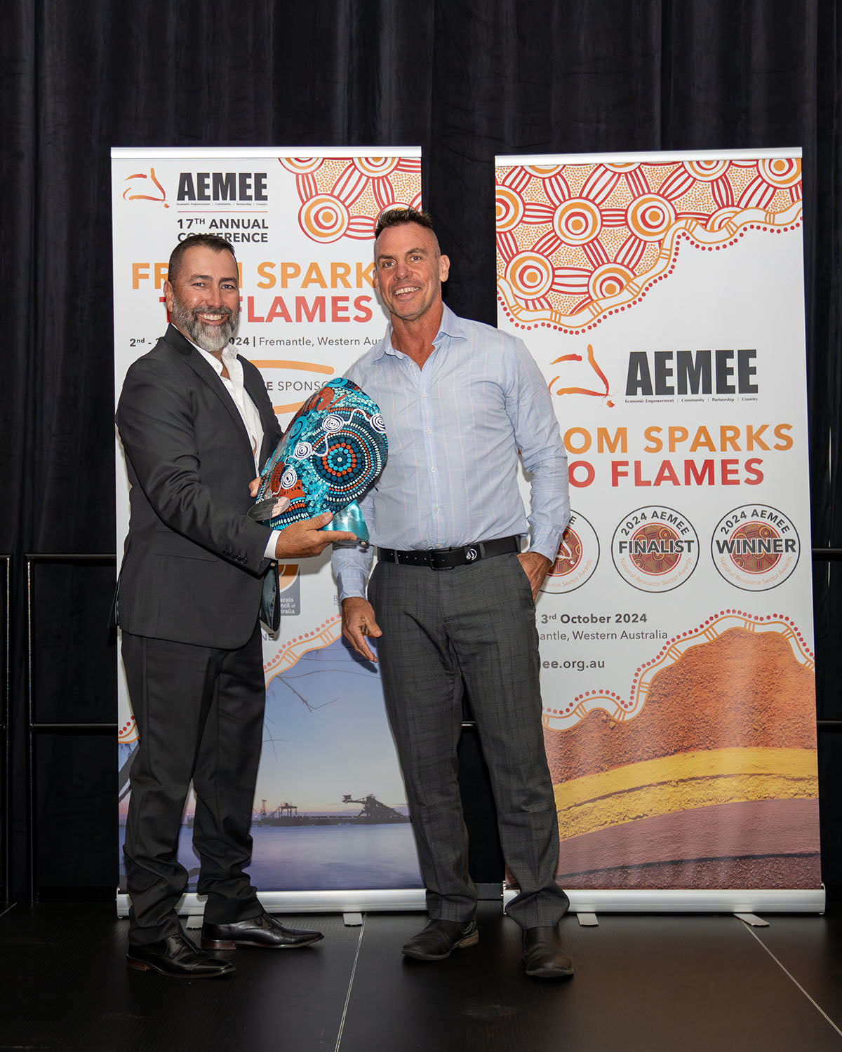AEMEE Chairperson  Derek Flucker smiling broadly alongside the winning representative from Biodiversity Australia and QGC, who is holding up their custom green and blue hand-painted oval trophy plaque for 2024 Resource Sector Collaboration of the Year.