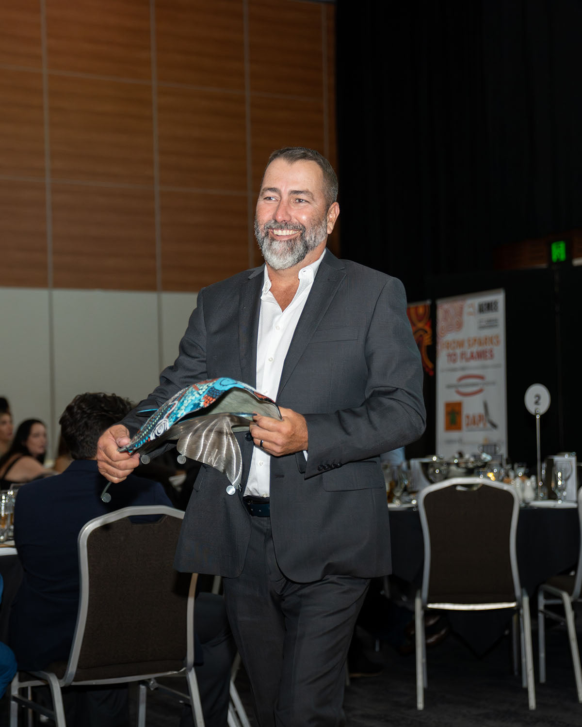 A close-up portrait of tRepresentatives from Waru Vehicle Hire smiling together on stage while holding their custom hand-painted dot-art award shield for 2024 New Indigenous Business of the Year in front of the official media banners.he representative from Biodiversity Australia and QGC smiling warmly while holding their custom hand-painted 2024 Resource Sector Collaboration of the Year award shield.