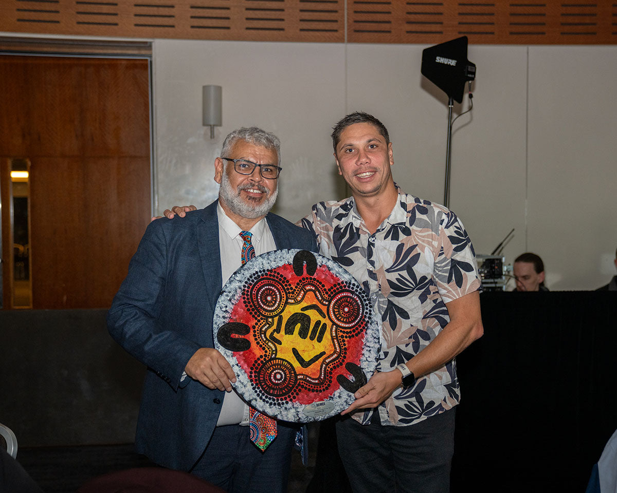 AEMEE CEO Jyi Lawton standing on stage with an official corporate event sponsor representative as they jointly showcase a beautifully vibrant, circular hand-painted Indigenous artwork trophy.