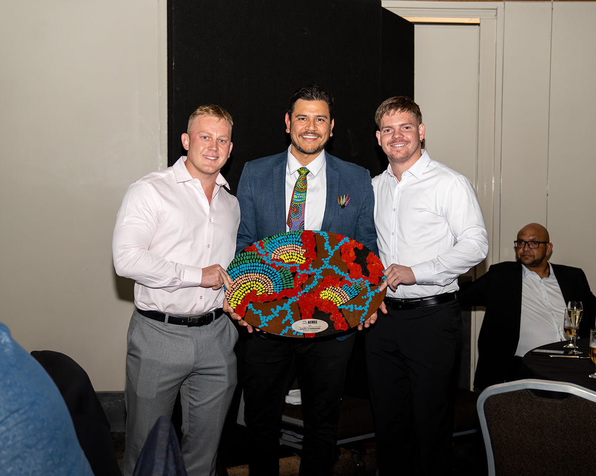 AEMEE Board Member and Dice Australia Director Raymond Pratt standing with two delegates on stage, smiling as they present a large, colorful hand-painted First Nations dot-art trophy shield.