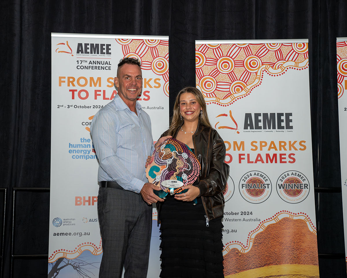 AMEE Director Derek Flucker, Chairperson and Representative from Waru Vehicle Hire smiling together on stage while holding their custom hand-painted dot-art award shield for 2024 New Indigenous Business of the Year in front of the official media banners.