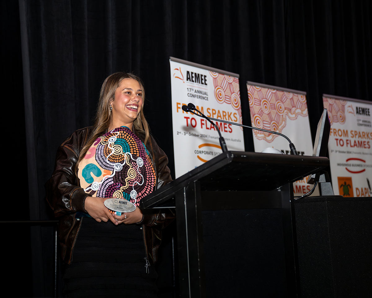 A female representative from Waru Vehicle Hire standing proudly behind the stage podium with her custom-painted Indigenous art trophy during her acceptance speech.