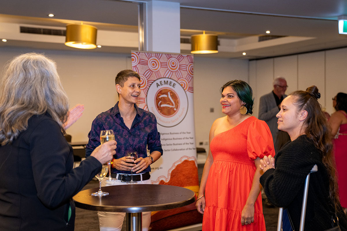 Delegates chatting and laughing around a high bar table with glasses of champagne during the pre-dinner mixer in front of an AEMEE banner.