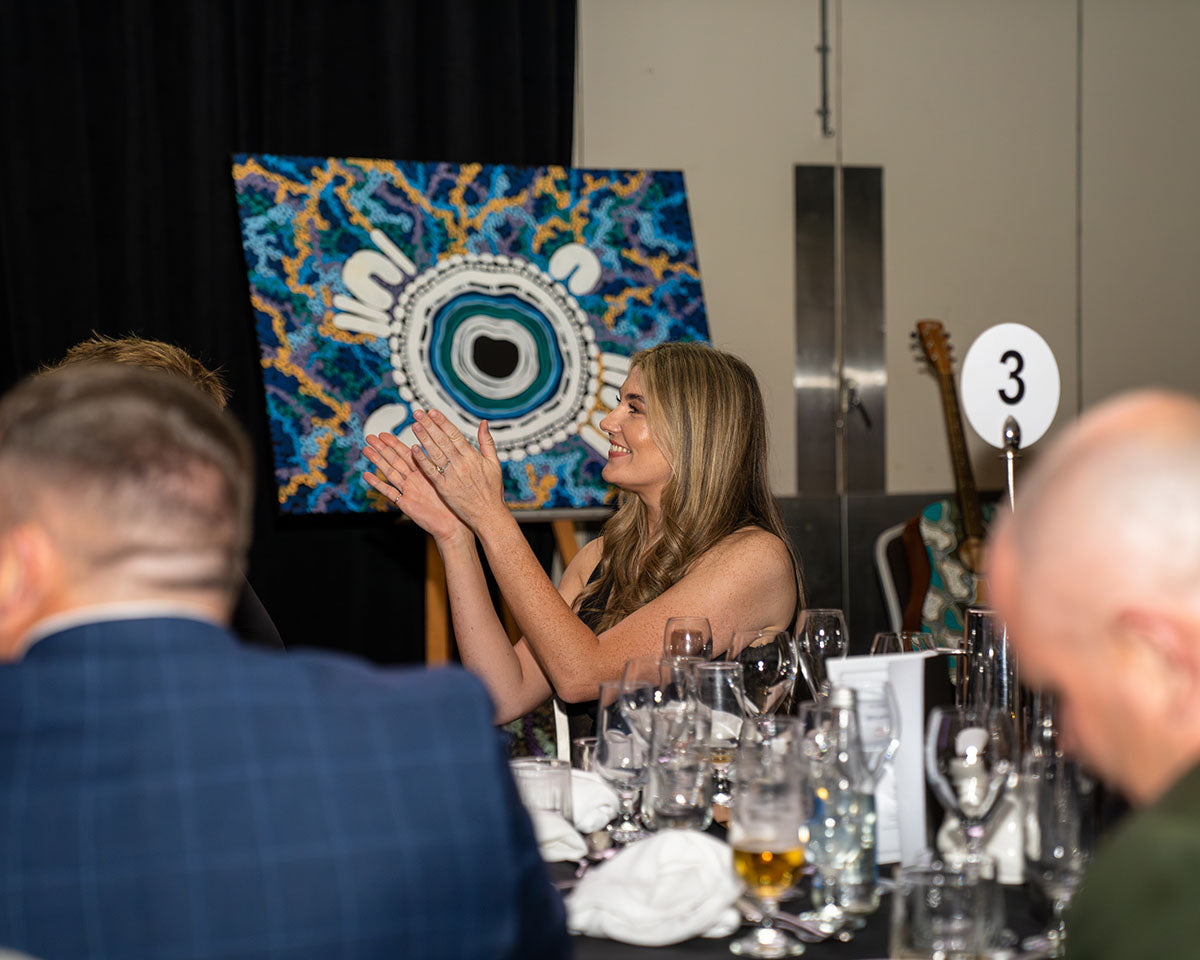 A female delegate smiling and applauding at table number three, with a premium hand-painted Indigenous artwork canvas on an easel visible in the background.