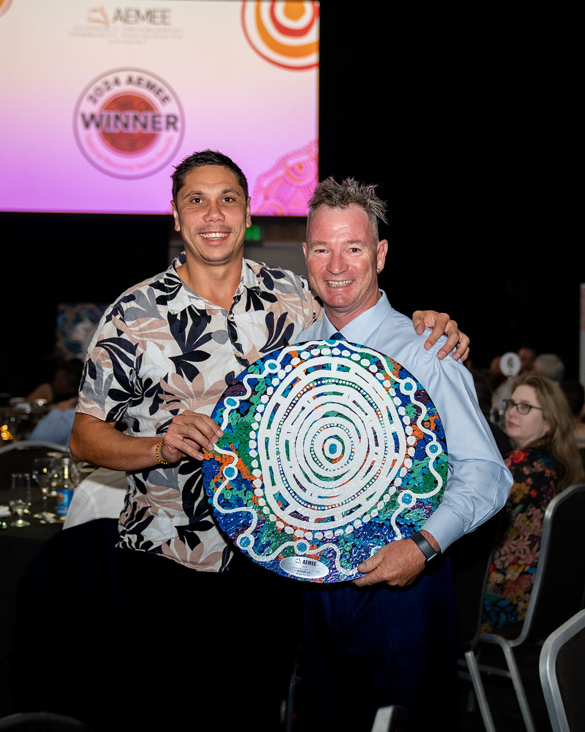 AEMEE CEO Jyi Lawton standing side-by-side with an executive winner, smiling warmly while holding up a large, intricate blue and white circular hand-painted Indigenous artwork award trophy.