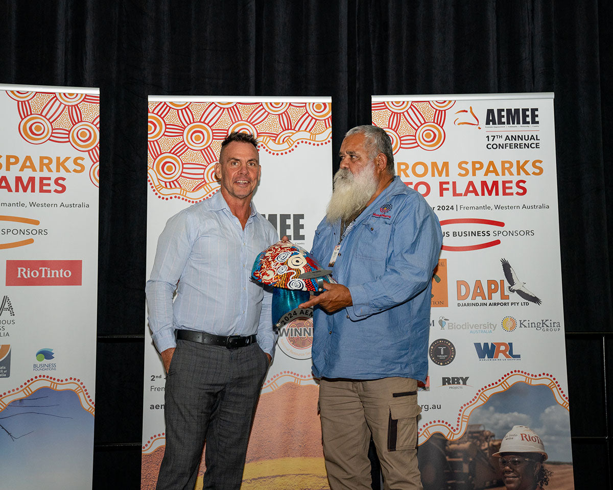 Derek Flucker standing on stage next to an Elder, presenting a custom hand-painted First Nations dot-art trophy shield in front of the official "From Sparks to Flames" media wall.