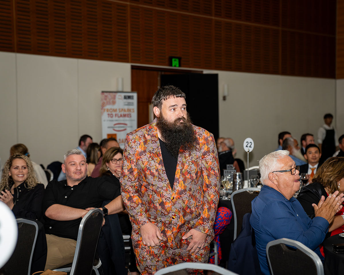 A representative from BlackRock Industries wearing a patterned orange blazer standing up from his dinner table to accept the 2024 Judges Choice award as guests applaud.