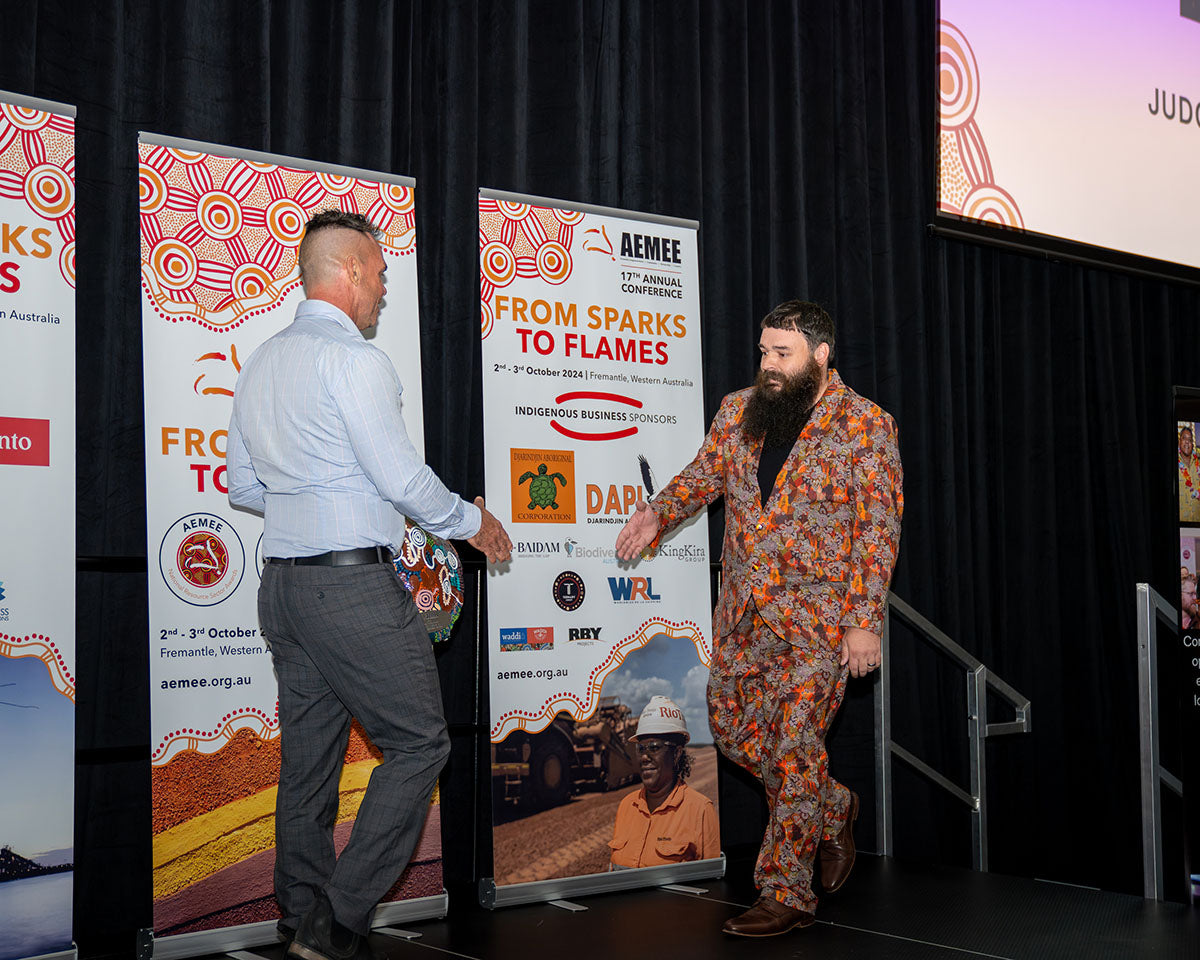 The representative from BlackRock Industries wearing a patterned orange blazer walking up onto the stage to accept the 2024 Judges Choice award in front of the official AEMEE conference media banners.