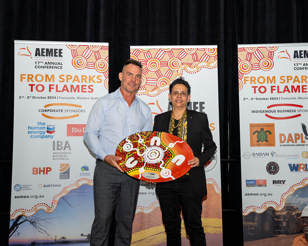 Tammy O'Connor standing on stage with a presenter, smiling proudly while showcasing a large, stunning orange and red custom hand-painted dot-art trophy shield for 2024 Sector Champion of the Year.