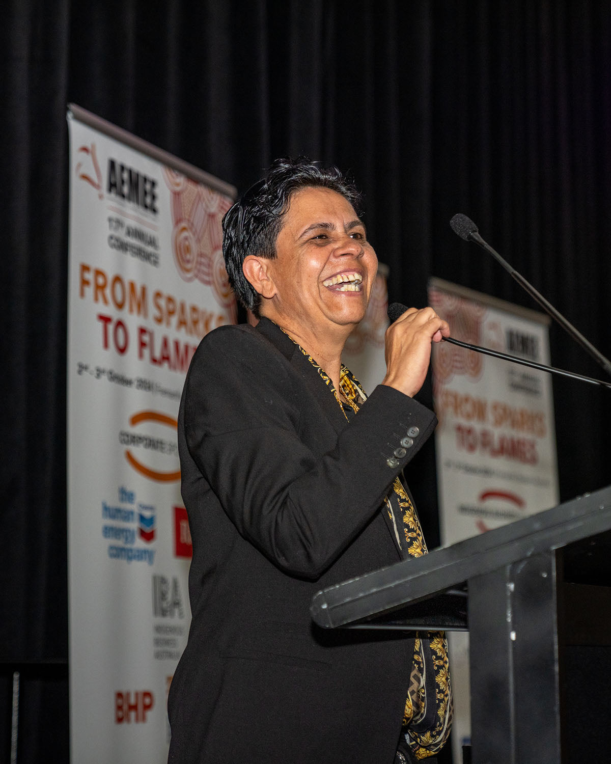 A close-up profile view of Tammy O'Connor laughing happily while speaking into the stage microphone during her acceptance speech for the 2024 Sector Champion of the Year award.