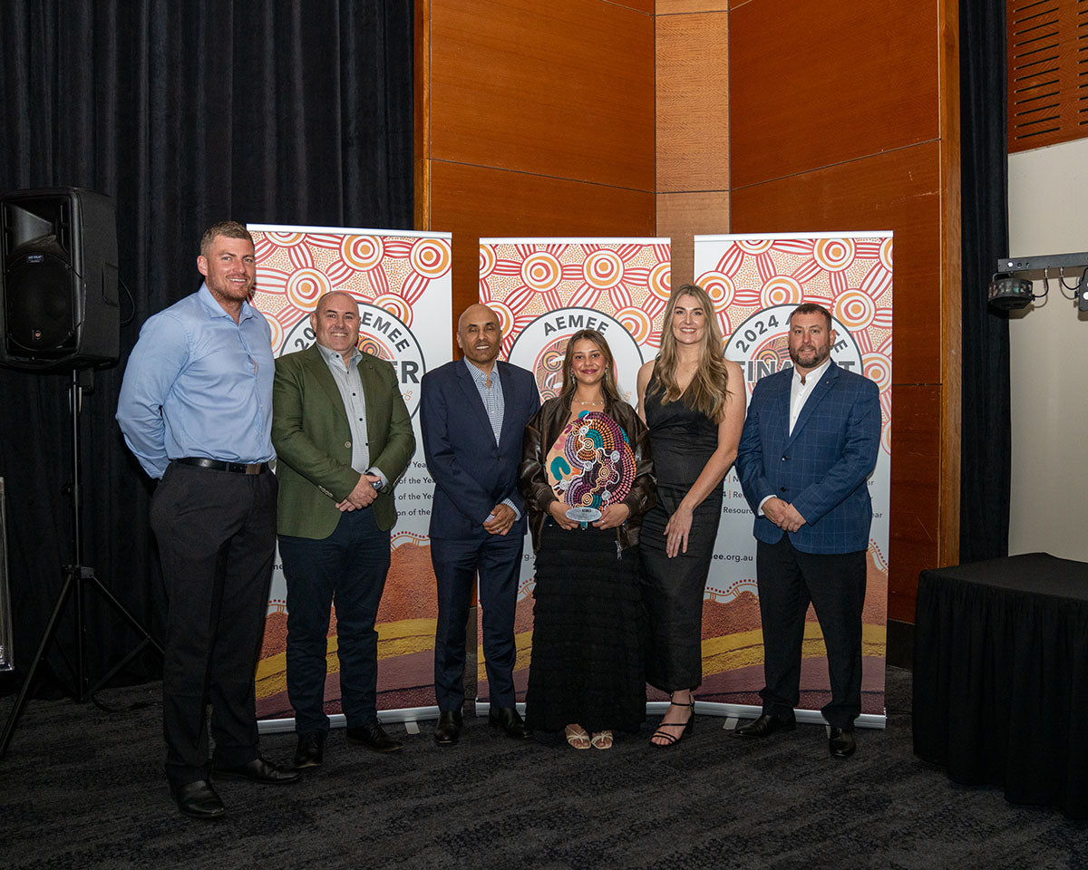 The corporate delegation from Waru Vehicle Hire posing together on stage, smiling proudly while showcasing their custom hand-painted dot-art award shield for 2024 New Indigenous Business of the Year.