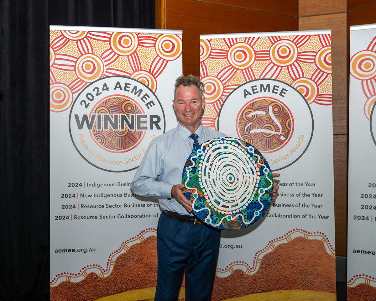 A winning corporate representative posing alone against the official 2024 winner banner while proudly holding a large, intricately patterned circular hand-painted Indigenous art trophy.