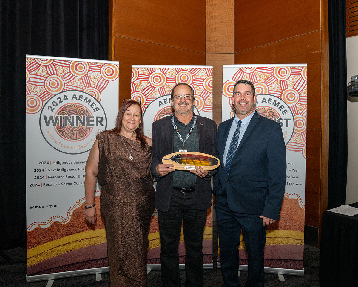 Roger Barnes standing between presenters on stage, smiling warmly while receiving his custom elongated hand-painted wooden trophy plaque for the 2024 Lifetime Supporter Award.