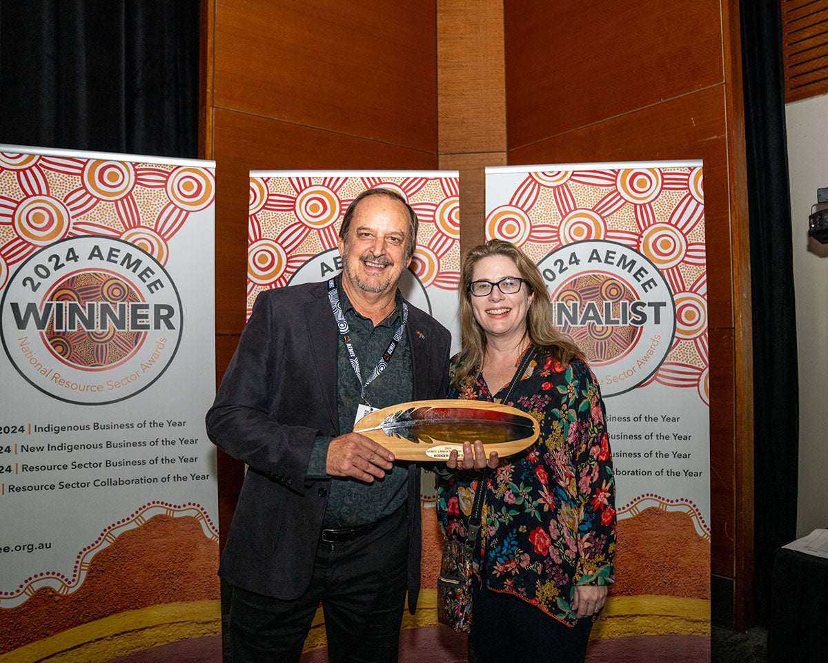 Roger Barnes posing with a presenter against the official 2024 Winner banner, holding up his custom-carved and hand-painted 2024 Lifetime Supporter Award plaque.