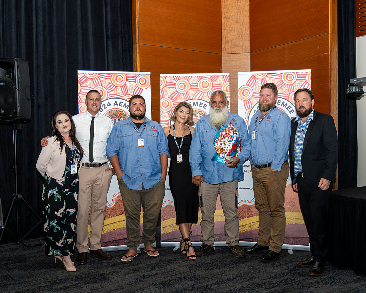 The MundaMurra team and an Elder gathered together on stage, standing proudly with their vibrant, custom hand-painted trophy shield to celebrate winning the 2024 Indigenous Business of the Year award.