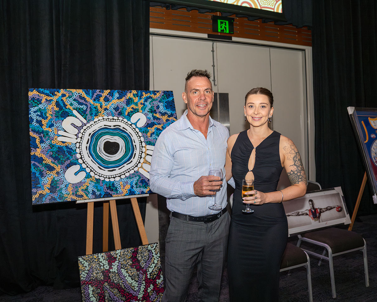 Derek Flucker and a female delegate holding drinks and smiling together inside the venue next to a display easel featuring a premium hand-painted Indigenous dot-art canvas.
