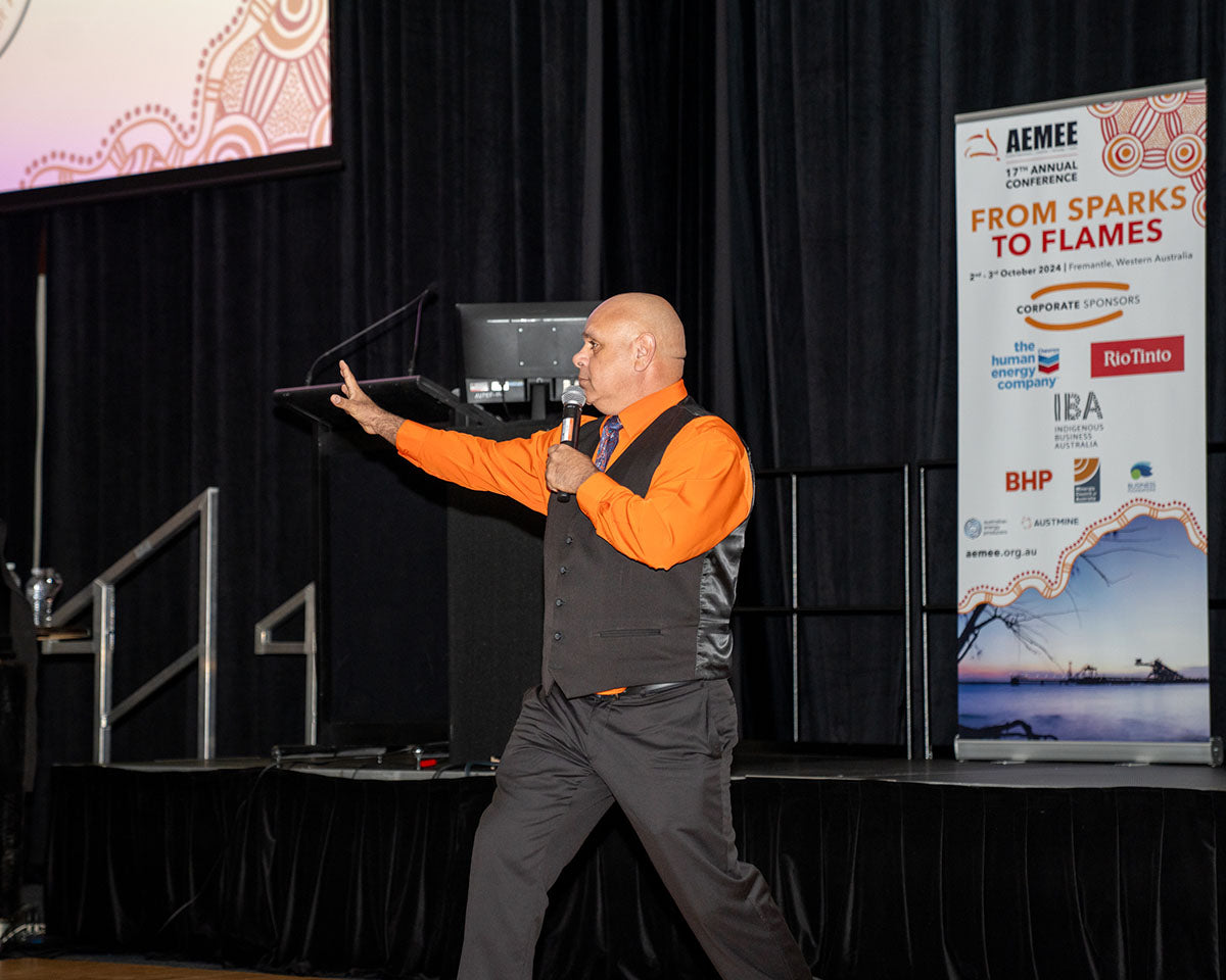 Comedian Kevin Kropinyeri performing live on stage in an orange shirt and black vest, gesturing dynamically with his arm outstretched while holding a microphone next to the official AEMEE "From Sparks to Flames" corporate sponsor banner.