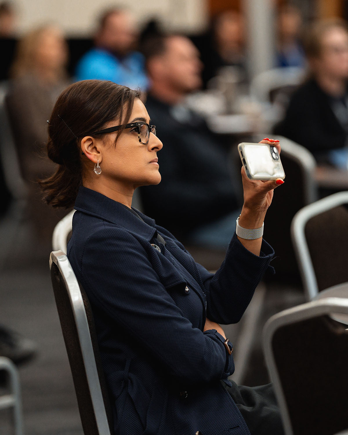 A female corporate delegate wearing glasses holding up her smartphone to record a keynote presentation from her table during the annual national AEMEE conference.