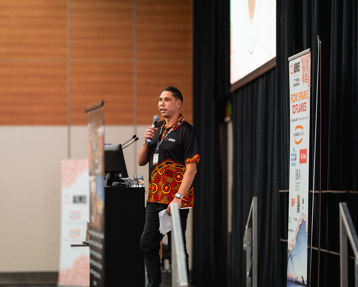 AEMEE CEO Jyi Lawton delivering an address into a microphone on stage, backdropped by corporate media banners at the annual national conference.