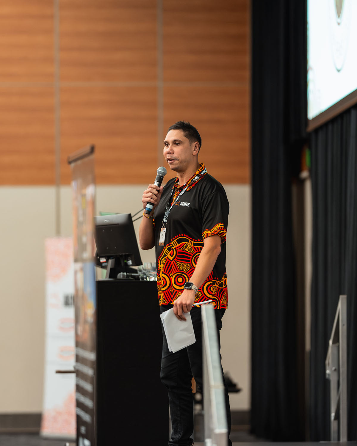 A medium profile shot of Jyi Lawton speaking at a microphone while wearing a traditional black and red artwork shirt during the annual AEMEE conference.