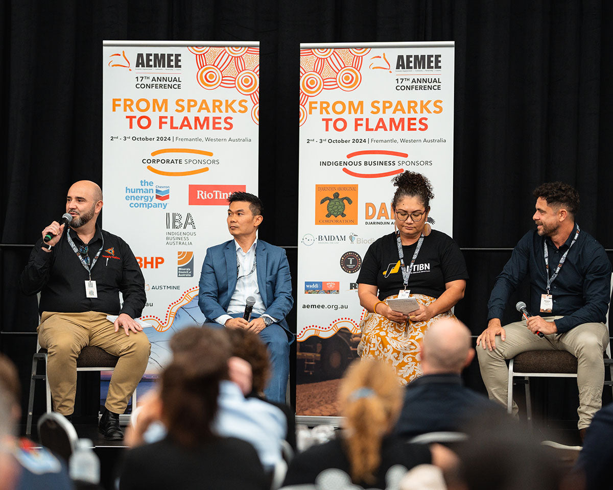 Seated presenters speaking to an audience on stage in front of official "From Sparks to Flames" corporate sponsor banners at the AEMEE conference.