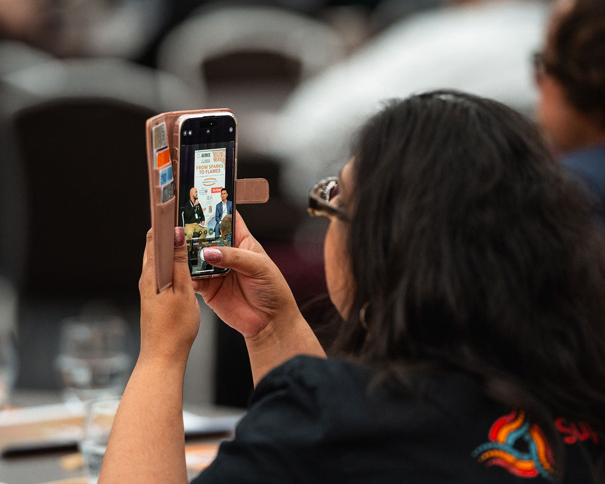A close-up view of a female delegate holding up her smartphone to record a live keynote presentation from her table during the AEMEE conference.