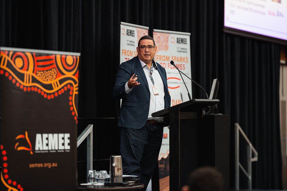A male corporate speaker in a suit jacket addressing conference delegates from the stage podium beside a traditional First Nations media banner.