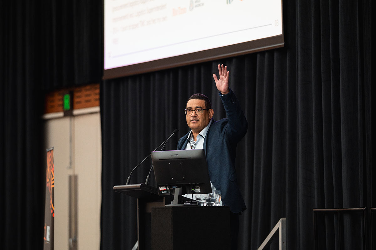A male presenter with his hand raised addressing audience questions beneath a projection screen at the annual national AEMEE conference.