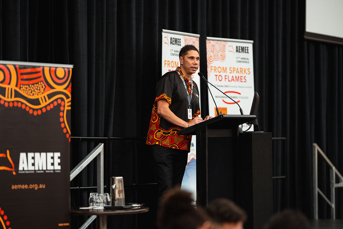 AEMEE CEO Jyi Lawton presenting from a side stage podium during a closing session at the AEMEE conference.