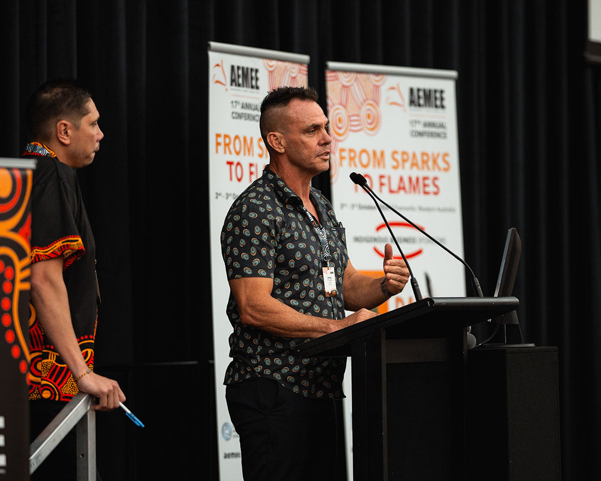 Chairperson Derek Flucker speaking at the microphone with hand gestures while AEMEE CEO Jyi Lawton stands nearby on the main stage.
