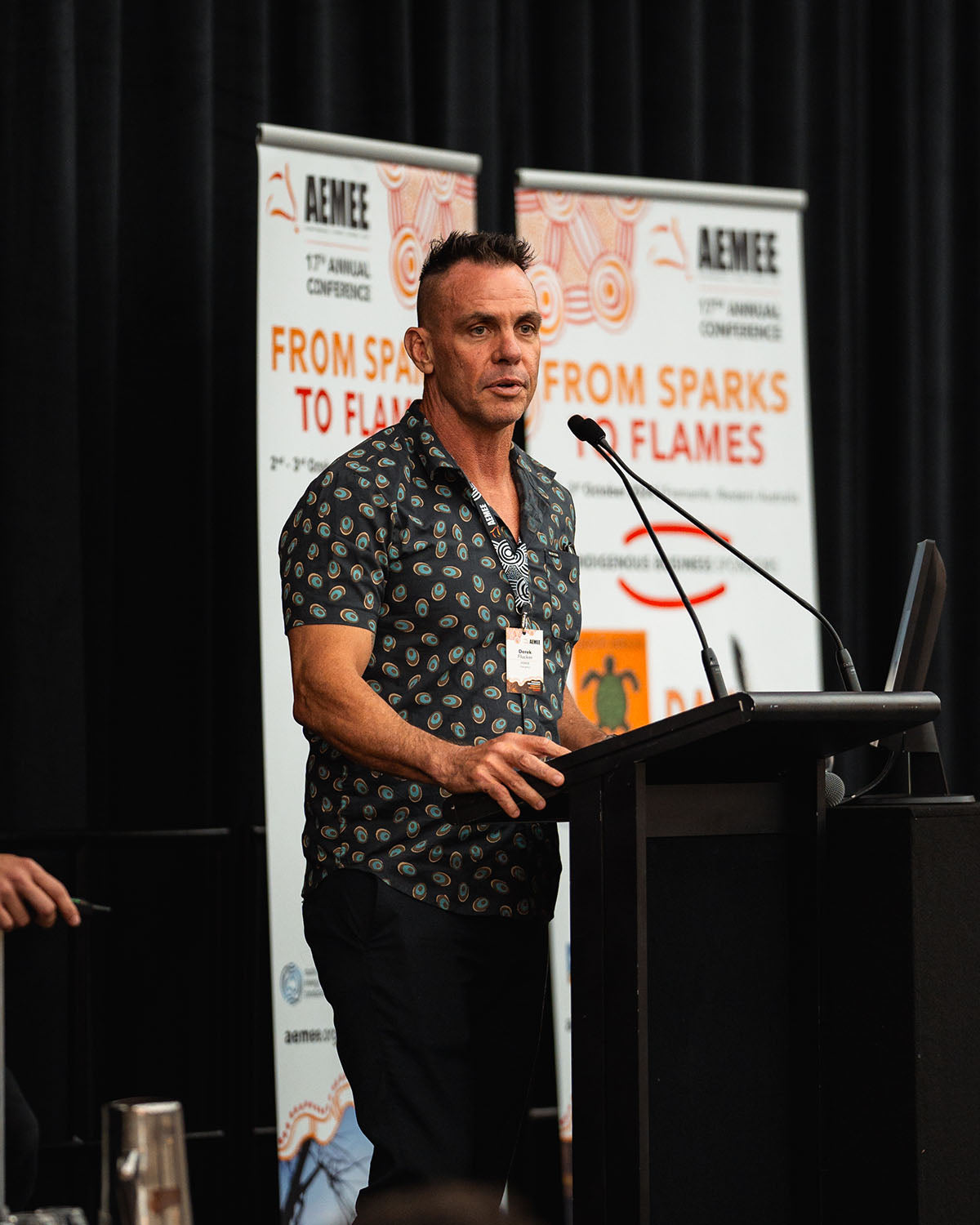 A close-up profile shot of AEMEE Chairperson Derek Flucker in a patterned collared shirt addressing delegates from behind the main podium.