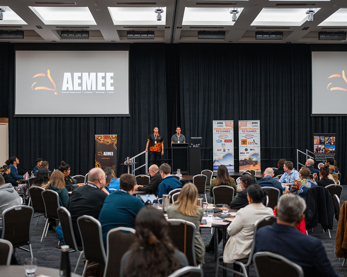 A wide viewpoint of seated conference delegates watching the leadership team present on stage beneath a large overhead project screen displaying the AEMEE logo.