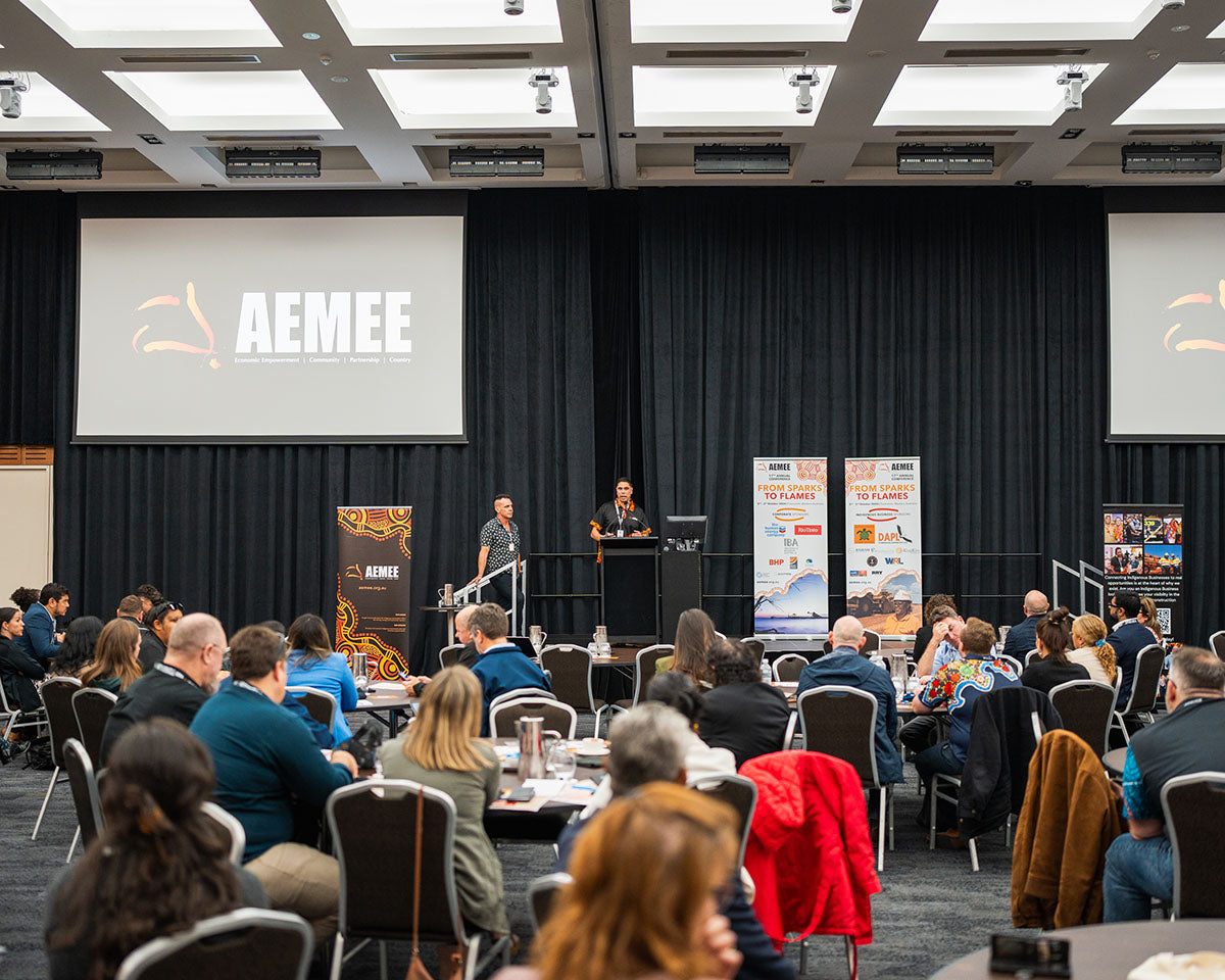 A wide view across rows of interactive tables filled with business professionals watching an executive address inside the main hall during the annual national AEMEE conference.